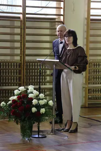 Obchody Powiatowego Dnia Edukacji Narodowej w sali gimnastycznej szkoły w Bełżycach: wystąpienia przy mównicy, wręczanie dyplomów i upominków nauczycielom oraz uczniom, goście na widowni, poczet sztandarowy i występ muzyczny uczniów.