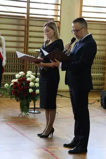Obchody Powiatowego Dnia Edukacji Narodowej w sali gimnastycznej szkoły w Bełżycach: wystąpienia przy mównicy, wręczanie dyplomów i upominków nauczycielom oraz uczniom, goście na widowni, poczet sztandarowy i występ muzyczny uczniów.