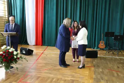 Obchody Powiatowego Dnia Edukacji Narodowej w sali gimnastycznej szkoły w Bełżycach: wystąpienia przy mównicy, wręczanie dyplomów i upominków nauczycielom oraz uczniom, goście na widowni, poczet sztandarowy i występ muzyczny uczniów.