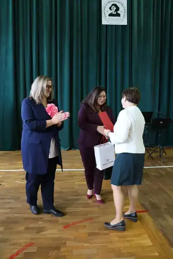 Obchody Powiatowego Dnia Edukacji Narodowej w sali gimnastycznej szkoły w Bełżycach: wystąpienia przy mównicy, wręczanie dyplomów i upominków nauczycielom oraz uczniom, goście na widowni, poczet sztandarowy i występ muzyczny uczniów.