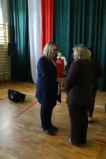 Obchody Powiatowego Dnia Edukacji Narodowej w sali gimnastycznej szkoły w Bełżycach: wystąpienia przy mównicy, wręczanie dyplomów i upominków nauczycielom oraz uczniom, goście na widowni, poczet sztandarowy i występ muzyczny uczniów.