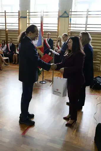 Obchody Powiatowego Dnia Edukacji Narodowej w sali gimnastycznej szkoły w Bełżycach: wystąpienia przy mównicy, wręczanie dyplomów i upominków nauczycielom oraz uczniom, goście na widowni, poczet sztandarowy i występ muzyczny uczniów.
