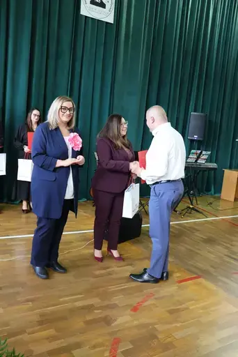 Obchody Powiatowego Dnia Edukacji Narodowej w sali gimnastycznej szkoły w Bełżycach: wystąpienia przy mównicy, wręczanie dyplomów i upominków nauczycielom oraz uczniom, goście na widowni, poczet sztandarowy i występ muzyczny uczniów.