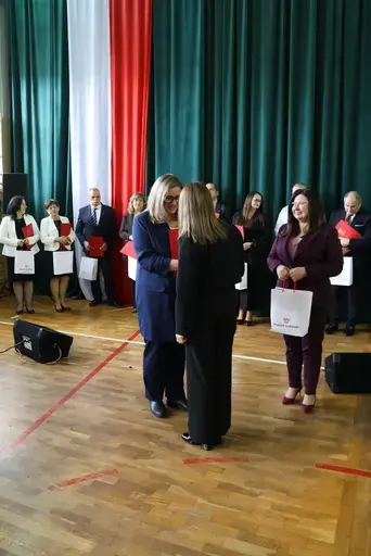 Obchody Powiatowego Dnia Edukacji Narodowej w sali gimnastycznej szkoły w Bełżycach: wystąpienia przy mównicy, wręczanie dyplomów i upominków nauczycielom oraz uczniom, goście na widowni, poczet sztandarowy i występ muzyczny uczniów.