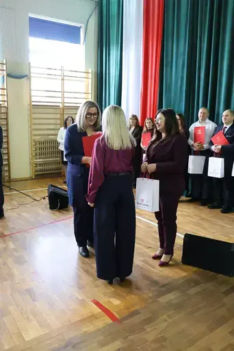 Obchody Powiatowego Dnia Edukacji Narodowej w sali gimnastycznej szkoły w Bełżycach: wystąpienia przy mównicy, wręczanie dyplomów i upominków nauczycielom oraz uczniom, goście na widowni, poczet sztandarowy i występ muzyczny uczniów.