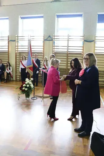 Obchody Powiatowego Dnia Edukacji Narodowej w sali gimnastycznej szkoły w Bełżycach: wystąpienia przy mównicy, wręczanie dyplomów i upominków nauczycielom oraz uczniom, goście na widowni, poczet sztandarowy i występ muzyczny uczniów.