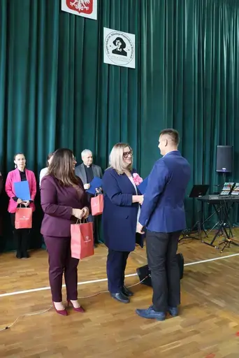 Obchody Powiatowego Dnia Edukacji Narodowej w sali gimnastycznej szkoły w Bełżycach: wystąpienia przy mównicy, wręczanie dyplomów i upominków nauczycielom oraz uczniom, goście na widowni, poczet sztandarowy i występ muzyczny uczniów.