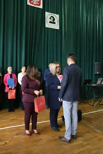 Obchody Powiatowego Dnia Edukacji Narodowej w sali gimnastycznej szkoły w Bełżycach: wystąpienia przy mównicy, wręczanie dyplomów i upominków nauczycielom oraz uczniom, goście na widowni, poczet sztandarowy i występ muzyczny uczniów.