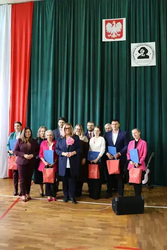 Obchody Powiatowego Dnia Edukacji Narodowej w sali gimnastycznej szkoły w Bełżycach: wystąpienia przy mównicy, wręczanie dyplomów i upominków nauczycielom oraz uczniom, goście na widowni, poczet sztandarowy i występ muzyczny uczniów.