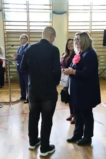 Obchody Powiatowego Dnia Edukacji Narodowej w sali gimnastycznej szkoły w Bełżycach: wystąpienia przy mównicy, wręczanie dyplomów i upominków nauczycielom oraz uczniom, goście na widowni, poczet sztandarowy i występ muzyczny uczniów.