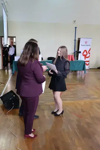 Obchody Powiatowego Dnia Edukacji Narodowej w sali gimnastycznej szkoły w Bełżycach: wystąpienia przy mównicy, wręczanie dyplomów i upominków nauczycielom oraz uczniom, goście na widowni, poczet sztandarowy i występ muzyczny uczniów.