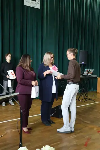 Obchody Powiatowego Dnia Edukacji Narodowej w sali gimnastycznej szkoły w Bełżycach: wystąpienia przy mównicy, wręczanie dyplomów i upominków nauczycielom oraz uczniom, goście na widowni, poczet sztandarowy i występ muzyczny uczniów.