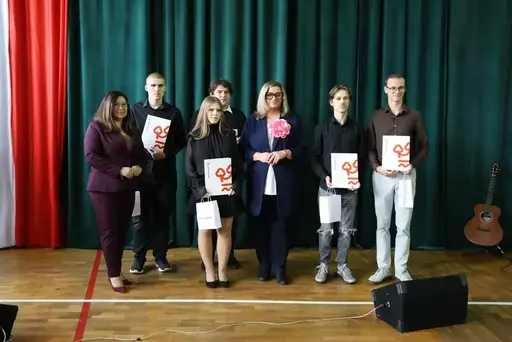 Obchody Powiatowego Dnia Edukacji Narodowej w sali gimnastycznej szkoły w Bełżycach: wystąpienia przy mównicy, wręczanie dyplomów i upominków nauczycielom oraz uczniom, goście na widowni, poczet sztandarowy i występ muzyczny uczniów.