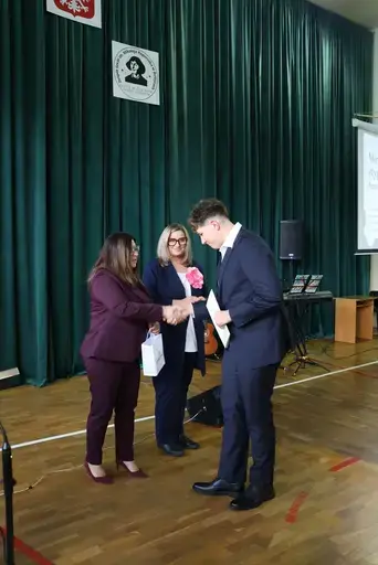 Obchody Powiatowego Dnia Edukacji Narodowej w sali gimnastycznej szkoły w Bełżycach: wystąpienia przy mównicy, wręczanie dyplomów i upominków nauczycielom oraz uczniom, goście na widowni, poczet sztandarowy i występ muzyczny uczniów.