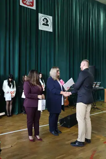 Obchody Powiatowego Dnia Edukacji Narodowej w sali gimnastycznej szkoły w Bełżycach: wystąpienia przy mównicy, wręczanie dyplomów i upominków nauczycielom oraz uczniom, goście na widowni, poczet sztandarowy i występ muzyczny uczniów.