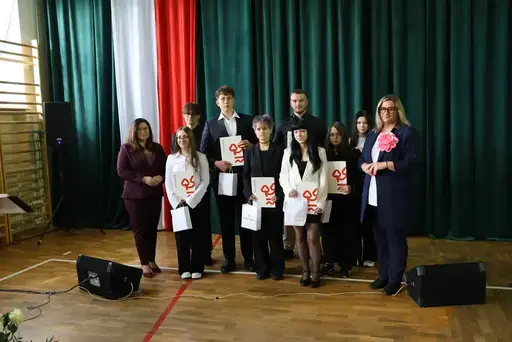 Obchody Powiatowego Dnia Edukacji Narodowej w sali gimnastycznej szkoły w Bełżycach: wystąpienia przy mównicy, wręczanie dyplomów i upominków nauczycielom oraz uczniom, goście na widowni, poczet sztandarowy i występ muzyczny uczniów.