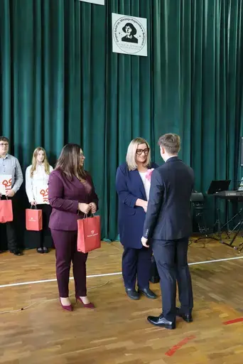 Obchody Powiatowego Dnia Edukacji Narodowej w sali gimnastycznej szkoły w Bełżycach: wystąpienia przy mównicy, wręczanie dyplomów i upominków nauczycielom oraz uczniom, goście na widowni, poczet sztandarowy i występ muzyczny uczniów.
