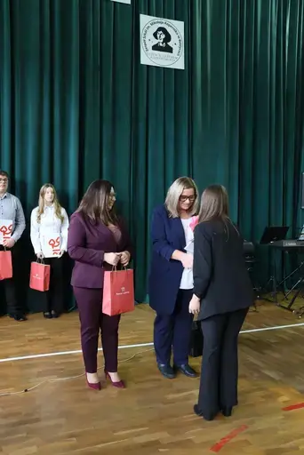 Obchody Powiatowego Dnia Edukacji Narodowej w sali gimnastycznej szkoły w Bełżycach: wystąpienia przy mównicy, wręczanie dyplomów i upominków nauczycielom oraz uczniom, goście na widowni, poczet sztandarowy i występ muzyczny uczniów.