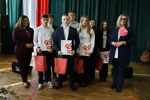 Obchody Powiatowego Dnia Edukacji Narodowej w sali gimnastycznej szkoły w Bełżycach: wystąpienia przy mównicy, wręczanie dyplomów i upominków nauczycielom oraz uczniom, goście na widowni, poczet sztandarowy i występ muzyczny uczniów.
