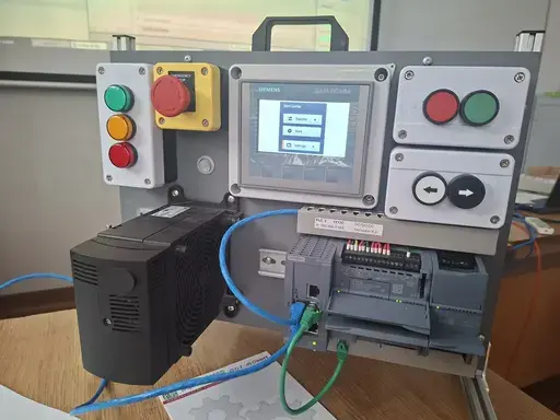 
                                                    Stanowisko dydaktyczne automatyki: metalowa płyta z panelem sterowniczym Siemens, przyciskami start-stop, czerwonym grzybkowym wyłącznikiem bezpieczeństwa w żółtej obudowie i kolorowymi lampkami. Poniżej zamontowany falownik i sterownik PLC z modułami, połączone niebieskim i zielonym przewodem sieciowym.
                                                