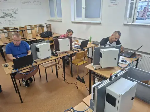 
                                                    Cztery osoby pracują przy laptopach w sali lekcyjnej. Każde stanowisko ma białą szafkę sterowniczą połączoną niebieskimi przewodami z komputerem. Na ścianie plansze techniczne, przy oknach ustawione zestawy ławek.
                                                