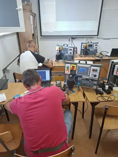 
                                                    Dwaj mężczyźni pracują przy stanowiskach ze sterownikami. Na pierwszym planie mężczyzna w bordowej koszulce siedzi tyłem przy laptopie i module sterowniczym z panelem operatorskim. W tle drugi uczestnik i duży ekran projekcyjny z wyświetlonym programem.
                                                