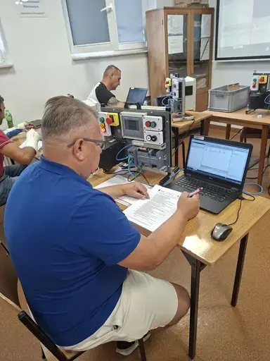 
                                                    Mężczyzna w niebieskiej koszulce polo i okularach robi notatki na wydrukowanych materiałach szkoleniowych. Obok stoi otwarty laptop z programem, a na sąsiednim stole zestaw sterowniczy z czerwonym wyłącznikiem bezpieczeństwa. W tle drugi uczestnik przy komputerze.
                                                