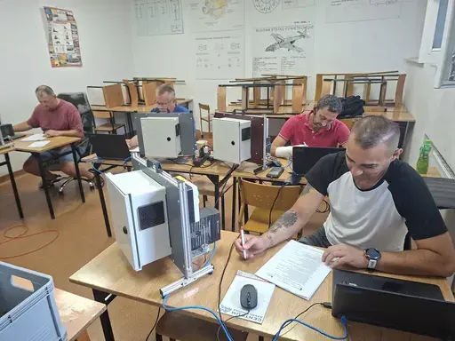 
                                                    Czterej mężczyźni siedzą przy ławkach ustawionych w rzędach i pracują na laptopach. Przy każdym stanowisku biała szafka sterownicza połączona przewodami. Na pierwszym planie uczestnik w jasnej koszulce wypełnia dokument długopisem.
                                                