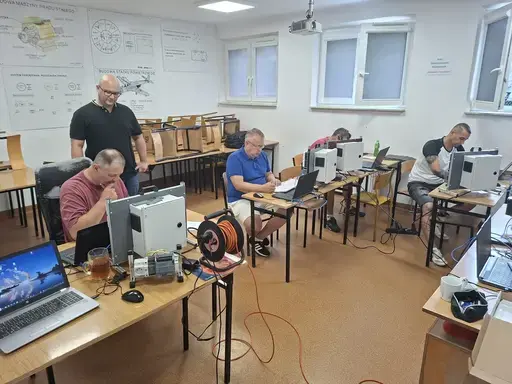 
                                                    Prowadzący w ciemnej koszulce stoi przy stanowisku jednego z uczestników i objaśnia zagadnienie. Pozostali czterej uczestnicy pracują przy laptopach i szafkach sterowniczych. Na podłodze pomarańczowy przedłużacz, na ścianach plansze techniczne.
                                                
