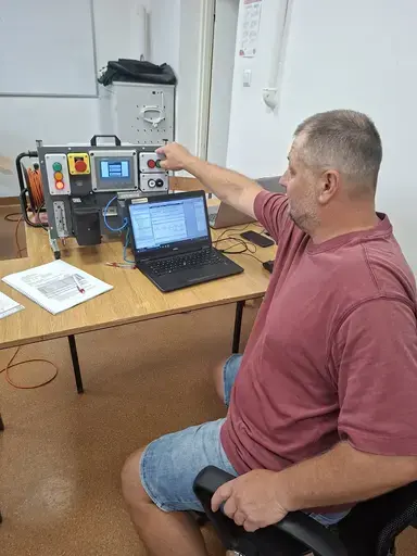 
                                                    Mężczyzna w bordowej koszulce siedzi przy stanowisku i wyciąga rękę w stronę panelu sterowniczego z wyświetlaczem i przyciskami. Obok otwarty laptop z uruchomionym programem, na stole materiały szkoleniowe.
                                                