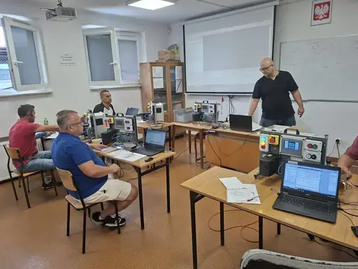 
                                                    Sala szkoleniowa: prowadzący w ciemnej koszulce stoi przy białej tablicy obok ekranu projekcyjnego i mówi do uczestników. Uczestnicy siedzą przy stołach z laptopami i zestawami sterowniczymi. Nad tablicą godło Polski.
                                                