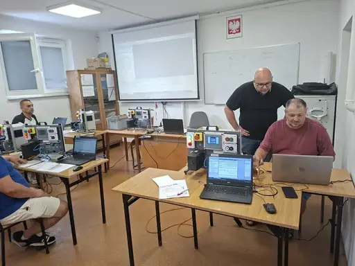 
                                                    Prowadzący w ciemnej koszulce pochyla się nad stanowiskiem uczestnika w bordowej koszulce i wskazuje coś na ekranie laptopa. Na stole dwa laptopy i moduł sterowniczy z panelem. W tle kolejni uczestnicy i ekran projekcyjny.
                                                