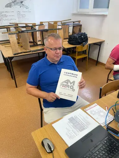 
                                                    Mężczyzna w niebieskiej koszulce polo i okularach siedzi przy stole i pokazuje zaświadczenie z logo Centrum Szkoleniowego AMPRO. Przed nim leżą materiały szkoleniowe i laptop. W tle poskładane ławki szkolne.
                                                