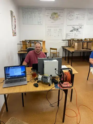 
                                                    Mężczyzna w bordowej koszulce siedzi przy stole naprzeciw stanowiska ze sterownikiem. Na blacie otwarty laptop z programem, silnik elektryczny i zwinięty pomarańczowy przewód. Na ścianie plansze z rysunkami technicznymi.
                                                