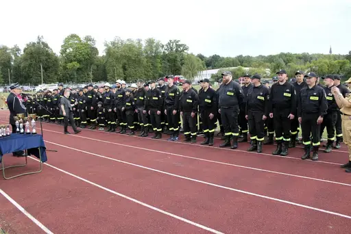 
                                                    Strażacy w ciemnych mundurach stoją w długim szyku wzdłuż czerwonej bieżni stadionu. Po lewej oficer w mundurze galowym, przed nim stolik nakryty granatowym obrusem z rzędem pucharów i statuetek. W tle drzewa, wozy strażackie i publiczność.
                                                