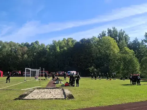 
                                                    Panorama trawiastego boiska otoczonego lasem w słoneczny dzień. Na murawie grupy strażaków w ciemnych mundurach, rozłożony sprzęt, bramka piłkarska i piaskownica skoczni. W głębi rząd czerwonych wozów strażackich i karetka.
                                                