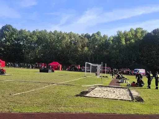 
                                                    Trawiaste boisko z rozciągniętymi wężami strażackimi i wyznaczonymi liniami toru. Po lewej czerwone namioty i stanowisko sędziowskie, w głębi czerwone wozy strażackie oraz grupy strażaków oczekujących na start. Tło stanowi ściana lasu.
                                                