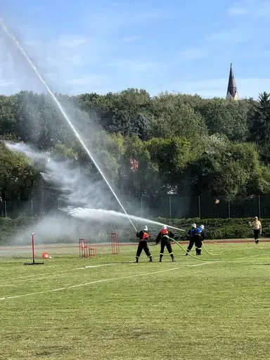 
                                                    Trzej strażacy w ciemnych mundurach i hełmach kierują prądownicę, z której w górę wystrzeliwuje długi strumień wody rozpylający się w mgłę. Przed nimi czerwone tarcze celownicze na stojakach. W tle drzewa i wieża kościoła.
                                                