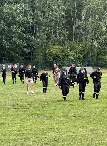 
                                                    Grupa strażaków i strażaczek w ciemnych mundurach z odblaskowymi pasami schodzi z trawiastego boiska po zakończonej konkurencji. Część idzie w czapkach z daszkiem, z tyłu ogrodzenie i drzewa.
                                                
