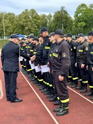 
                                                    Oficer w granatowym mundurze galowym podaje rękę i wręcza dyplomy strażakom ustawionym w szpalerze na czerwonej bieżni. Strażacy w ciemnych mundurach z naszywkami Straż trzymają w rękach białe dyplomy.
                                                