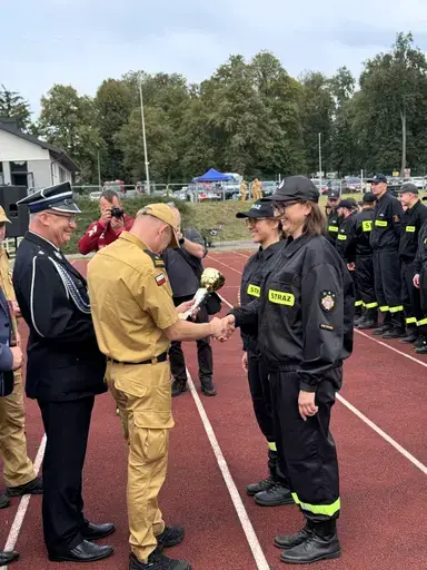 
                                                    Oficer w piaskowym mundurze wręcza złoty puchar uśmiechniętej strażaczce w ciemnym mundurze z naszywką Straż. Obok stoi druga strażaczka i oficer w mundurze galowym, w tle fotograf i szpaler uczestników na bieżni.
                                                