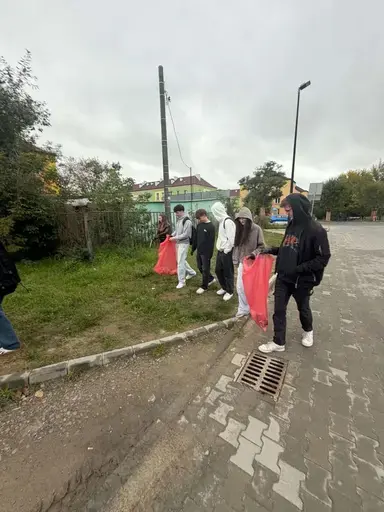 
                                                    Uczniowie Zespołu Szkół Zawodowych nr 2 w Dęblinie w małych grupach zbierają odpady do niebieskich i czerwonych worków na trawnikach i chodnikach z kostki wzdłuż osiedlowych ulic, w tle bloki, pawilony handlowe i drzewa.
                                                