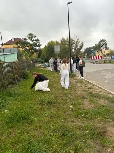 
                                                    Uczniowie Zespołu Szkół Zawodowych nr 2 w Dęblinie w małych grupach zbierają odpady do niebieskich i czerwonych worków na trawnikach i chodnikach z kostki wzdłuż osiedlowych ulic, w tle bloki, pawilony handlowe i drzewa.
                                                