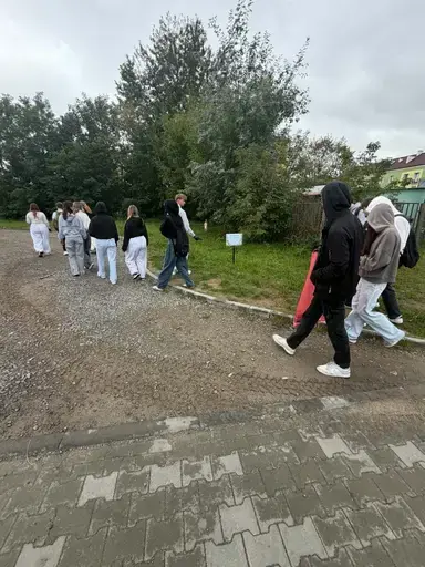 
                                                    Uczniowie Zespołu Szkół Zawodowych nr 2 w Dęblinie w małych grupach zbierają odpady do niebieskich i czerwonych worków na trawnikach i chodnikach z kostki wzdłuż osiedlowych ulic, w tle bloki, pawilony handlowe i drzewa.
                                                