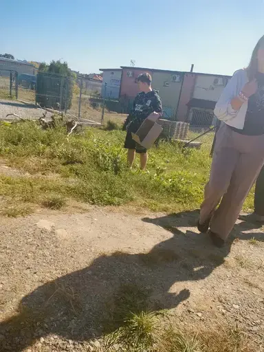 
                                                    Uczniowie Zespołu Szkół Zawodowych nr 2 w Dęblinie w małych grupach zbierają odpady do niebieskich i czerwonych worków na trawnikach i chodnikach z kostki wzdłuż osiedlowych ulic, w tle bloki, pawilony handlowe i drzewa.
                                                