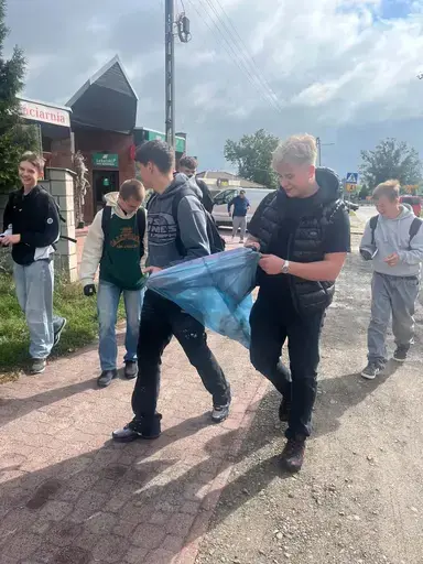
                                                    Uczniowie Zespołu Szkół Zawodowych nr 2 w Dęblinie w małych grupach zbierają odpady do niebieskich i czerwonych worków na trawnikach i chodnikach z kostki wzdłuż osiedlowych ulic, w tle bloki, pawilony handlowe i drzewa.
                                                