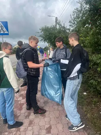 
                                                    Uczniowie Zespołu Szkół Zawodowych nr 2 w Dęblinie w małych grupach zbierają odpady do niebieskich i czerwonych worków na trawnikach i chodnikach z kostki wzdłuż osiedlowych ulic, w tle bloki, pawilony handlowe i drzewa.
                                                