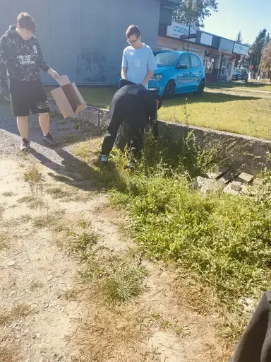 
                                                    Uczniowie Zespołu Szkół Zawodowych nr 2 w Dęblinie w małych grupach zbierają odpady do niebieskich i czerwonych worków na trawnikach i chodnikach z kostki wzdłuż osiedlowych ulic, w tle bloki, pawilony handlowe i drzewa.
                                                