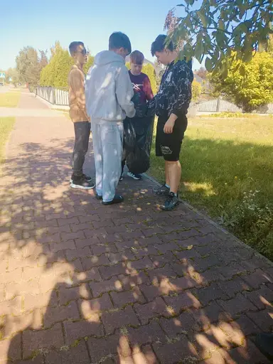 
                                                    Uczniowie Zespołu Szkół Zawodowych nr 2 w Dęblinie w małych grupach zbierają odpady do niebieskich i czerwonych worków na trawnikach i chodnikach z kostki wzdłuż osiedlowych ulic, w tle bloki, pawilony handlowe i drzewa.
                                                