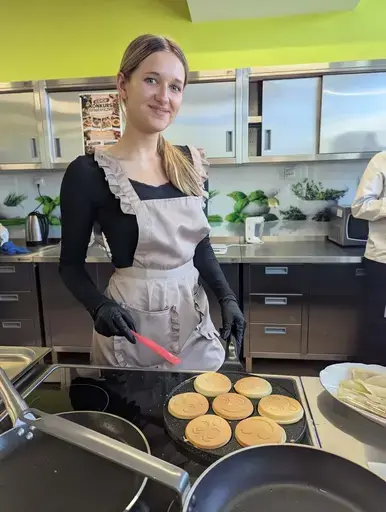 
                                                    Uczniowie Zespołu Szkół Zawodowych nr 2 w Dęblinie w fartuchach smażą i nakładają naleśniki w szkolnej pracowni gastronomicznej oraz przy stoiskach nakrytych zielonymi obrusami, z czarnymi banerami z nazwami dań, udekorowanych różowymi i białymi balonami; na stołach porcje naleśników z dodatkami na białych talerzykach.
                                                
